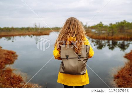 Woman traveler in a yellow coat walks along a wooden walking path along the wetlands. Back view. Woman traveler in a yellow coat walks along a wooden walking path along the wetlands. Back view. 117667948