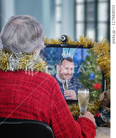 Cheerful, Connected Christmas Eve: A Senior Woman in a Festive Red Sweater Sits at Her Desk, Engaged in a Video Call with a Man Sporting a Bunny Hat Who Raises a Glass of Sparkling Wine. Cheerful, Connected Christmas Eve: A Senior Woman in a Festive Red Sweater Sits at Her Desk, Engaged in a Video Call with a Man Sporting a Bunny Hat Who Raises a Glass of Sparkling Wine. 117668032