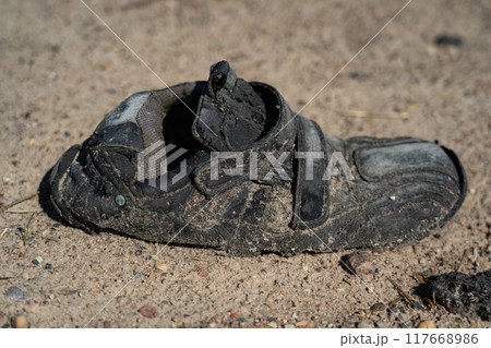 An old, dirty, wrinkled, black shoe, lying on a gravel road 117668986