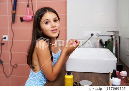 Young girl applying lip balm in a bathroom with a sink and hair dryer 117669595
