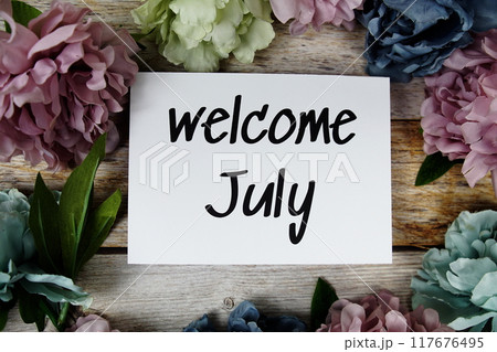 Welcome July text on paper card with flowers frame top view on wooden background 117676495