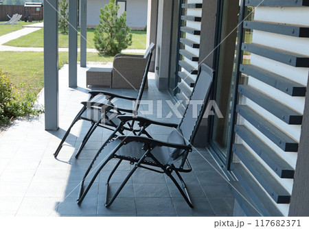 Flexible folding patio chairs in shade under awning of modern house in summer. Garden furniture and relaxation. Furniture design. Flexible folding patio chairs in shade under awning of modern house in summer. Garden furniture and relaxation. Furniture design. 117682371