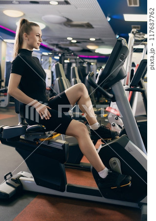 Young woman exercising on a stationary bike in a modern gym during daytime 117687762