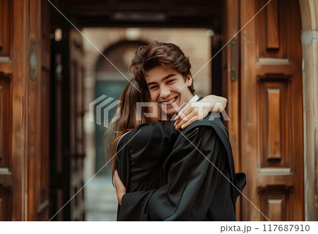 A young man wearing a black graduation gown and cap is posing with his girlfriend stand in front of a university's corridor, showing love for each other, look happy after just having graduated A young man wearing a black graduation gown and cap is posing with his girlfriend stand in front of a university's corridor, showing love for each other, look happy after just having graduated 117687910