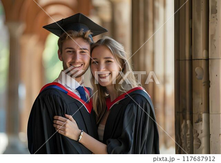 A young man wearing a black graduation gown and cap is posing with his girlfriend stand in front of a university's corridor, showing love for each other, look happy after just having graduated 117687912