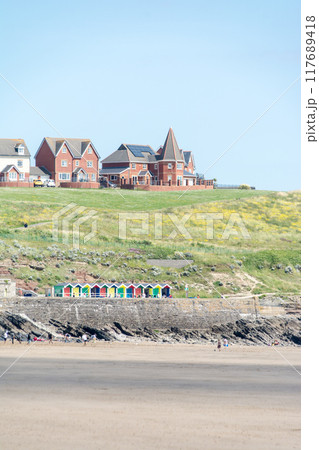 遠浅の砂浜の海岸でくつろぐ人々と遠くの丘の上に立つ赤煉瓦の家並み　南ウェールズの郊外にて 117689418