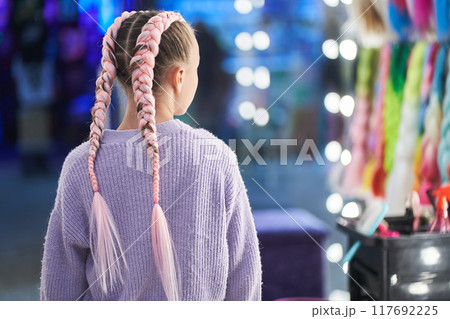 An unrecognizable girl with pigtails made of pink kanekalon. The girl looks at herself in the mirror and is delighted with her new hairstyle from the kanekalon in the beauty salon. High quality photo 117692225