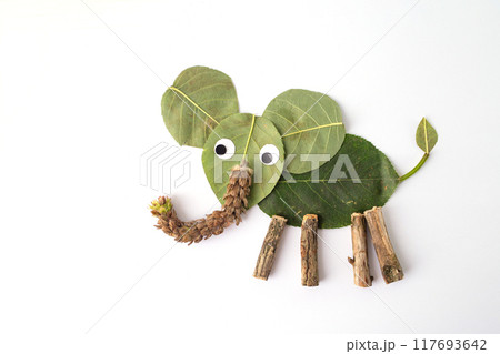 A creative craft project where natural elements like leaves, twigs, and a seed pod form an elephant. Large leaf body, smaller leaf ears, white eyes with black centers, seed pod trunk, twig legs 117693642