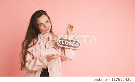Upset young woman with closed sign in pink studio Upset young woman with closed sign in pink studio 117695420
