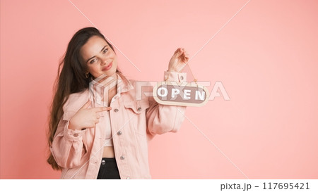 Smiling woman with open sign inviting in pink studio Smiling woman with open sign inviting in pink studio 117695421