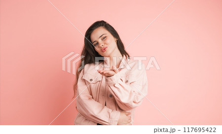 Happy woman blowing kiss and showing heart gesture in pink studio 117695422