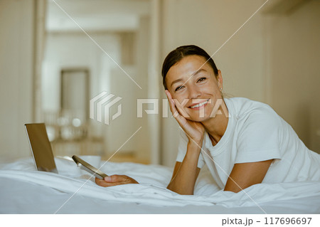 A young woman relaxes in her cozy bed, engaged with a laptop and phone, enjoying leisure A young woman relaxes in her cozy bed, engaged with a laptop and phone, enjoying leisure 117696697