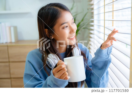 Young asian woman standing beside window and holding mug in bedroom at home 117701580