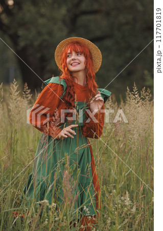 A shepherdess woman with red hair and a green dress sits in a field of flowers. A shepherdess woman with red hair and a green dress sits in a field of flowers. 117701819