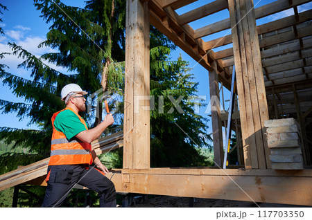 Carpenter constructing wooden frame house near the forest. Bearded man hammering nails into structure while wearing protective helmet and construction vest. Concept of modern ecological construction. Carpenter constructing wooden frame house near the forest. Bearded man hammering nails into structure while wearing protective helmet and construction vest. Concept of modern ecological construction. 117703350