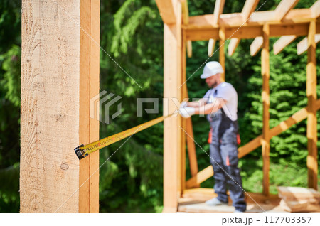 Carpenter building wooden-framed home. Selective focus of man measures distances with measuring tape while wearing work clothes and helmet. Concept of contemporary, eco-friendly building practices. Carpenter building wooden-framed home. Selective focus of man measures distances with measuring tape while wearing work clothes and helmet. Concept of contemporary, eco-friendly building practices. 117703357