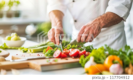 A chef preparing fresh vegetable salad in a bright kitchen. The chef slices tomatoes along with other fresh ingredients on a wooden cutting board. 117704912