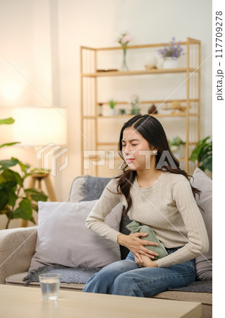 A woman is sitting on a couch with a bottle of water and a heating pad 117709278