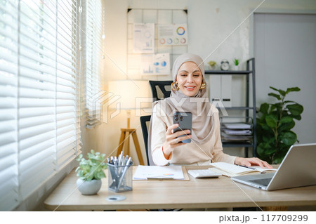 A woman wearing a hijab is sitting at a desk with a laptop and a cell phone A woman wearing a hijab is sitting at a desk with a laptop and a cell phone 117709299