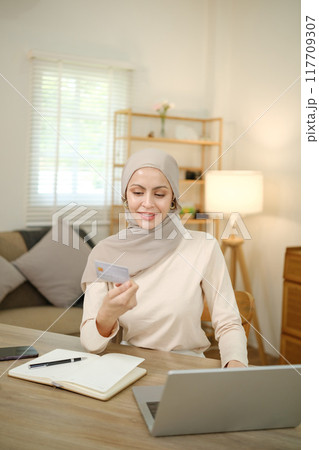 A woman wearing a head scarf is sitting at a desk with a laptop and a notebook 117709307