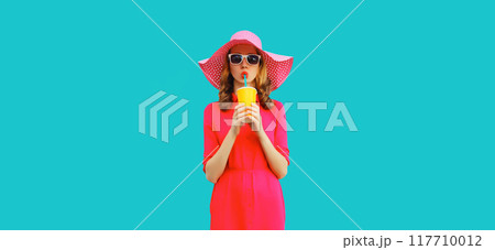 Summer portrait of beautiful young woman drinking fresh juice in hat, pink dress on blue background 117710012
