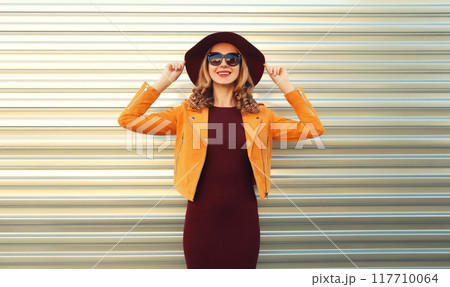 Beautiful smiling young woman wearing orange jacket, round hat on gray background 117710064
