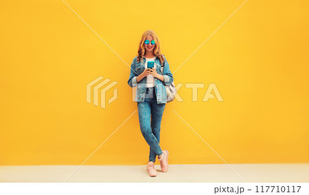 Portrait of stylish modern smiling young woman with smartphone in jean jacket on yellow background Portrait of stylish modern smiling young woman with smartphone in jean jacket on yellow background 117710117