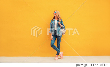 Full length beautiful smiling young woman wearing denim jacket, backpack on yellow background Full length beautiful smiling young woman wearing denim jacket, backpack on yellow background 117710118