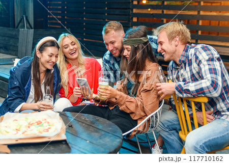Beautiful woman shows interesting stuff on her smartphone to her friends while they spend time together on a weekend party outdoors. 117710162