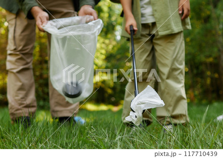 Two individuals picking up litter with trash grabber in wooded park area on sunny day, maintaining cleanliness and contributing to environmental conservation Two individuals picking up litter with trash grabber in wooded park area on sunny day, maintaining cleanliness and contributing to environmental conservation 117710439
