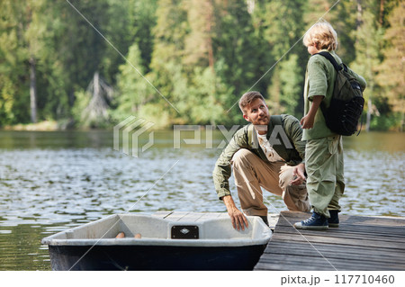 Father kneeling on dock while talking to son beside lake surrounded by trees, both dressed in casual outdoor clothes carrying fishing gear 117710460