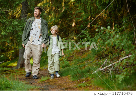 Father and son walking hand in hand along green forest trail on sunny day. Both dressed in casual hiking outfits, enjoying nature and each other's company 117710479