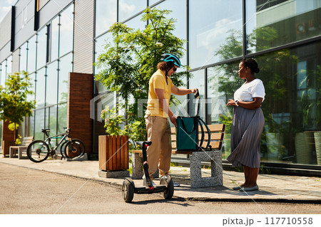 Delivering person on electric scooter handing package to woman waiting on bench with greenery and sunlight illuminating scene outside modern building 117710558