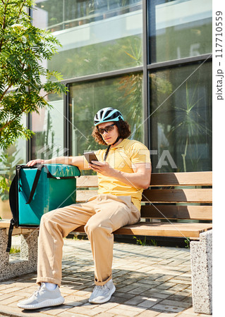 Cyclist with curly hair sitting on bench outside modern building and checking smartphone while resting during delivery work wearing helmet and casual clothing 117710559