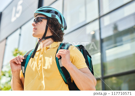 Outdoor cyclist preparing for ride, wearing helmet and sunglasses, looking away from camera while holding backpack straps 117710571