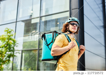 Bicyclist wearing yellow polo shirt and helmet, carrying teal insulated bag for food delivery and standing in front of glass building 117710572