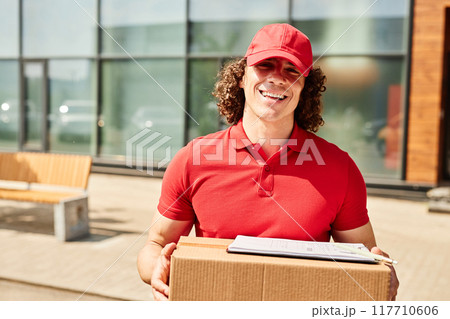 Smiling delivery worker holding package and clipboard while standing outside near building in bright sunny weather Smiling delivery worker holding package and clipboard while standing outside near building in bright sunny weather 117710606