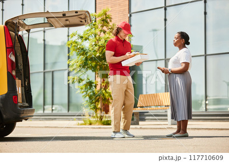 Delivery worker handing over package, with customer receiving it near delivery van. Scene depicts professional service interaction outside a commercial building Delivery worker handing over package, with customer receiving it near delivery van. Scene depicts professional service interaction outside a commercial building 117710609