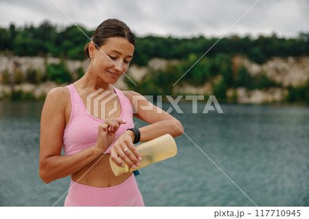 An Active Woman is Intently Checking Her Smartwatch While Enjoying the Scenic Water View An Active Woman is Intently Checking Her Smartwatch While Enjoying the Scenic Water View 117710945
