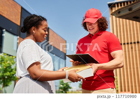 Smiling delivery person handing over package to cheerful customer outdoors near buildings on sunny day creating positive interaction Smiling delivery person handing over package to cheerful customer outdoors near buildings on sunny day creating positive interaction 117710980