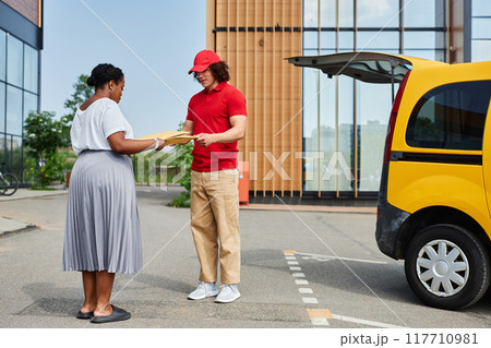 Courier in red cap and recipient standing near yellow van in sunny outdoors, holding package and engaging in conversation Courier in red cap and recipient standing near yellow van in sunny outdoors, holding package and engaging in conversation 117710981