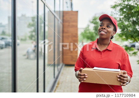 African American courier smiling while holding package Outdoors and wearing red uniform and cap person standing next to building with glass windows scene in background 117710984