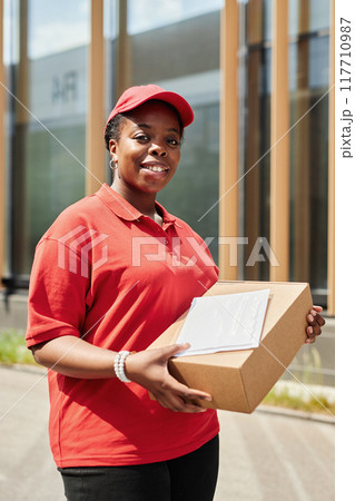 African American female worker holding package while standing outside of commercial building and smiling at camera. Wearing red uniform and cap, conveying sense of professionalism and friendliness 117710987