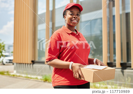 Delivery worker smiling while holding package in urban environment, wearing uniform with red cap and shirt, person standing outdoors next to building with sunlight illuminating 117710989