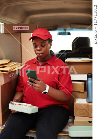 African American woman seated among packages and boxes while using mobile phone for updates on shipment status 117711012