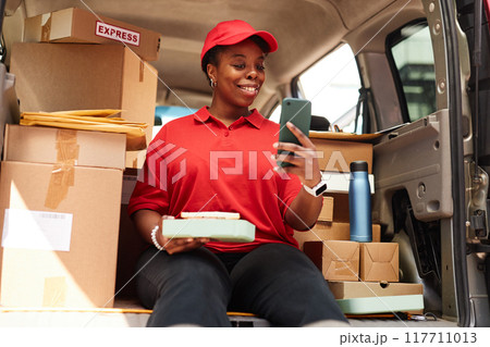 Smiling delivery worker holding package while looking at smartphone in delivery vehicle surrounded by boxes smiling and wearing red uniform 117711013