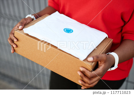 Person holding cardboard package with white label sticker on top wearing red shirt standing outside building 117711023
