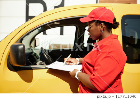 Delivery person in red uniform standing beside yellow vehicle while checking and signing delivery documents, reflecting busy work environment Delivery person in red uniform standing beside yellow vehicle while checking and signing delivery documents, reflecting busy work environment 117711040