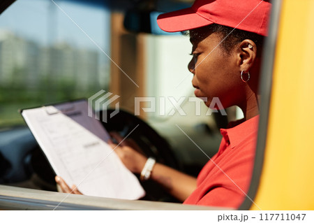 African American woman in red uniform reviewing checklist while sitting in delivery van with focus on task at hand and clipboard in hand. Background shows blurry urban setting 117711047