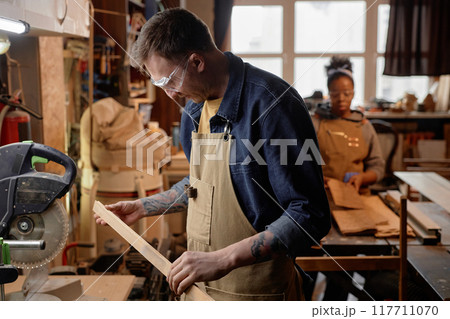 Man working in artisan workshop with focus on wooden piece and tools visible around him. Person in background also engaging in crafting activity in rustic wooden workshop 117711070